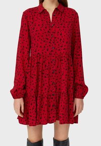 Robe florale rouge à manches longues, fermeture boutonnée sur le devant, taille froncée et jupe à volants. Le tissu est léger et texturé, avec un imprimé floral noir.