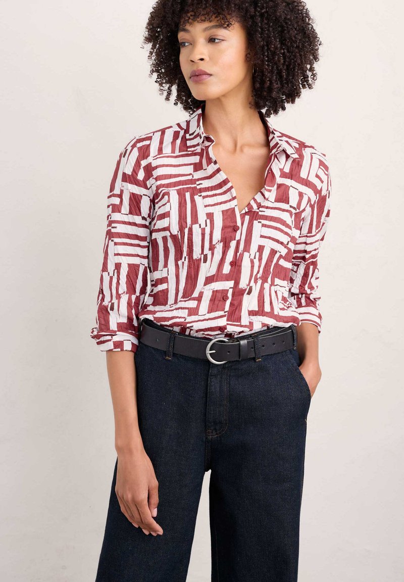 Camicia a righe rossa e bianca con colletto a V, indossata con pantaloni in denim scuri e una cintura nera con fibbia rotonda argentata.