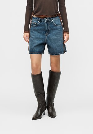 Person iført mørkeblå denimshorts, brun langærmet bluse og høje sorte støvler med hæl, stående på en ensfarvet baggrund.