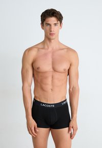 Schwarze Baumwoll-Boxershorts mit glatter Textur und elastischem Bund, der ein weißes "LACOSTE"-Logo trägt. Das Modell zeigt ein figurschmeichelndes Design.