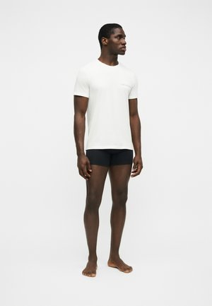 Homme debout pieds nus portant un t-shirt blanc à col rond et un boxer noir sur un fond blanc uni.