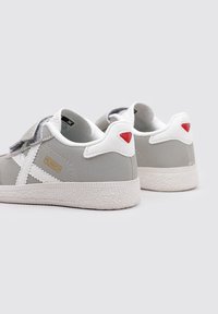 Zapatos de suede gris con acentos blancos, que cuentan con una suela texturizada blanca, una correa de Velcro y un detalle triangular rojo en el talón.