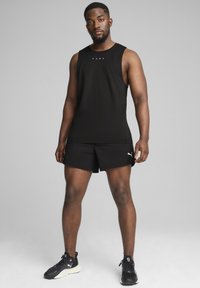 Camiseta atlética sin mangas negra con logo, combinada con pantalones cortos negros. El modelo lleva zapatillas negras. Textura suave, diseño ajustado.