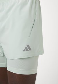 Gaiši zaļi sporta šorti ar vaļīgu ārējo kārtu un pieguļošām iekšējām šortiem. Funkcijas ietver ventilācijas caurumi un pelēks logo sānos.