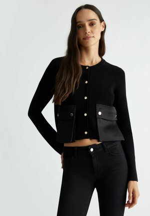Cardigan nero a coste con bottoni dorati, caratterizzato da due tasche frontali in materiale liscio. Design accorciato, abbinato a jeans neri.