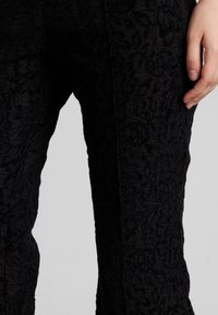 Pantalones de encaje negro con un patrón floral texturizado, un ajuste a medida y un acabado suave a lo largo de las piernas, capturados a mitad del muslo.