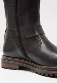 Bota de cuero negro hasta la mitad de la pantorrilla con cierre de cremallera lateral, suela plana y acentos texturizados. Presenta una suela exterior de goma marrón contrastante.