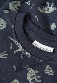 Felpa blu navy con motivo di dinosauri verdi e ossa, colletto e polsini a coste, e un'etichetta con la dicitura "basic needs" attaccata al colletto.