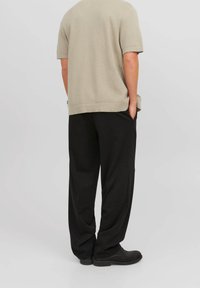 Pullover beige a maniche corte e texture, abbinato a pantaloni neri larghi e scarpe nere. Il modello sta in piedi con le mani in tasca, rivolto di spalle.