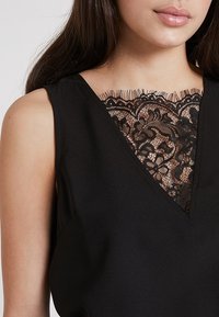 Svart ärmlös topp med V-halsdesign och intrikata svarta spetsdetaljer vid bröstet, slät tyg och en figursydd silhuett.