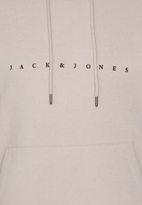 Šviesiai smėlinis džemperis, pagamintas iš minkštos medžiagos, su juodu spaudiniu "JACK & JONES" logotipu priekyje, reguliuojama stringų kapuce ir priekinė kišenė.