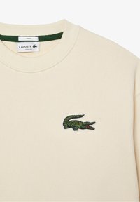 Kremowy sweter z prążkowanej tkaniny, z zielonym haftowanym logo krokodyla po lewej stronie klatki piersiowej oraz zielonym wykończeniem dekoltu.