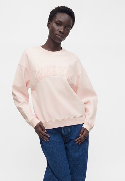 Γυναίκα με ανοιχτό ροζ oversized φούτερ με ανάγλυφο κείμενο και φαρδιά μπλε τζιν, στέκεται με τα χέρια κοντά στους γοφούς μπροστά σε λευκό φόντο.