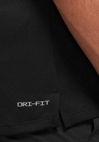 Gros plan sur un tissu noir avec le texte "DRI-FIT" imprimé près d'un petit ourlet fendu cousu et une partie du bras d'une personne.