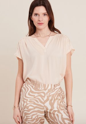 Blouse van zacht beige stof met een V-hals en korte mouwen, met haakwerkdetails op de schouders. Gecombineerd met beige broeken met zebra print.
