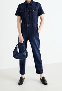 Combinaison en denim à manches courtes, avec deux poches poitrine et une taille cintrée. Associée à un sac à main texturé bleu foncé et des mocassins noirs.