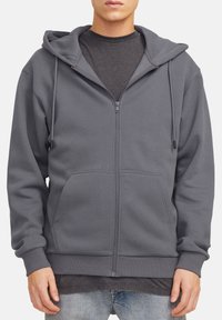 Sweat à capuche gris zippé avec cordon, poches avant et poignets côtelés. Fabriqué en tissu doux avec une texture lisse. Superposé sur une chemise sombre.