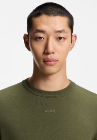 Olivengrøn strikket sweater med ribbet rund halsudskæring og et subtilt "HUGO"-logo på brystet. Glat tekstur, tætsiddende design.