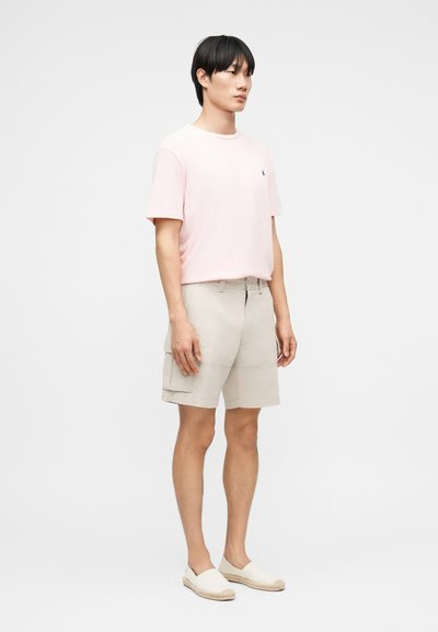 Νεαρός άνδρας που στέκεται πλαγίως, φοράει ροζ ανοιχτό T-shirt, μπεζ σορτς καρό και μπεζ κα slip-on παπούτσια, μπροστά σε απλό λευκό φόντο.