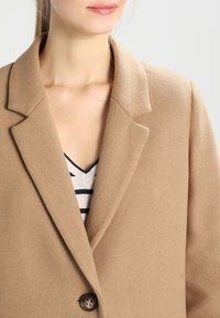 Blazer in misto lana di colore marrone chiaro con rever a punta, chiusura con singolo bottone e texture morbida, indossato sopra una maglietta a righe.