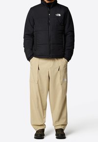 Chaqueta de plumas negra con cremallera frontal y cuello alzado, combinada con pantalones cargo beiges con bolsillos y un logo. Botas negras completan el conjunto.