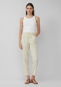 Witte geribde tanktop gecombineerd met lichtbeige, loszittende broek met zijzakken en een trekkoord in de taille. Bruine platte sandalen.