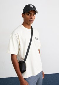 Cream short-sleeve t-shirt with black "Dickies" logo, worn with gray pants and a small black crossbody bag, set against a plain background.