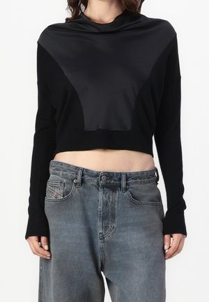 Femme portant un pull court noir avec un panneau avant triangulaire effet satin, associé à un jean gris délavé ample.
