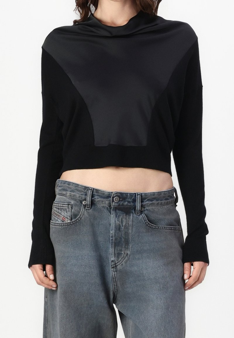 Femme portant un pull court noir avec un panneau avant triangulaire effet satin, associé à un jean gris délavé ample.