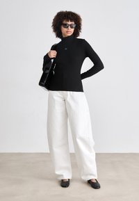 Zwarte lange mouw turtleneck top met een klein logo, gecombineerd met losse witte broek en zwarte platte schoenen. Model houdt een zwarte handtas vast.