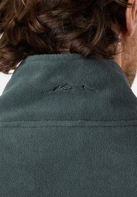 Tumma vihreä fleece-takki, jossa on korkea kaulus, takana on hienovarainen vuoristologon brodeeraus, pehmeä pintakuvio ja ei näkyviä kiinnityksiä.