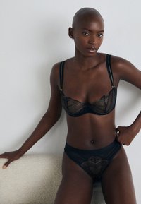 Conjunto de lingerie de renda preta com um sutiã com detalhes transparentes e acabamentos em dourado, combinado com uma thong a condizer, ambos apresentando padrões intricados.