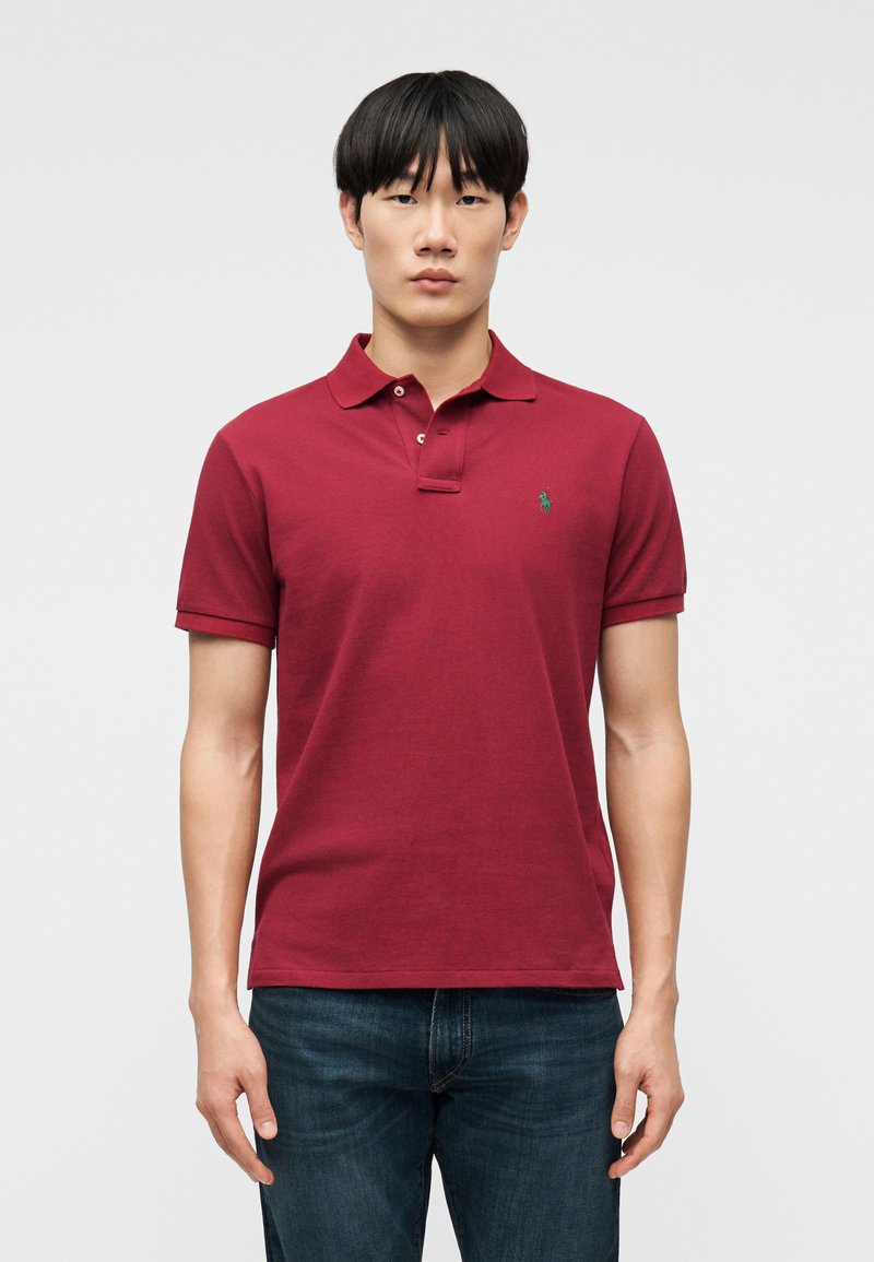 Νεαρός άνδρας που κοιτάζει μπροστά, φοράει κόκκινο πόλο με κοντά μανίκια και μικρό πράσινο λογότυπο, καθώς και σκούρο μπλε τζιν, μπροστά σε απλό φόντο.