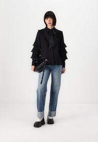 Top nero con maniche a volant, colletto legato. Jeans blu con risvolti. Borsa nera, scarpe nere chunky. Sfondo bianco semplice.