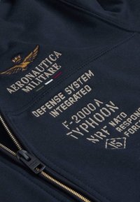 Giacca in tessuto blu navy con ricami dorati. Presenta una cerniera, un'insegna militare e il testo "Aeronautica Militare" e "Typhoon."