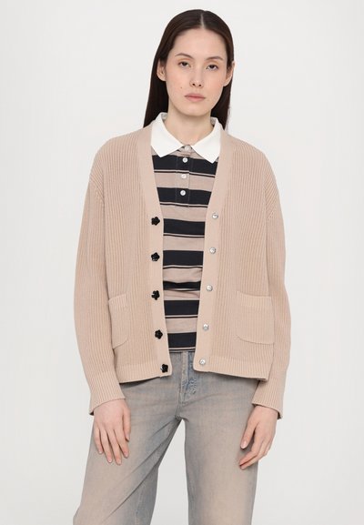 Μπεζ πλεκτό cardigan με V-λαιμό, μπροστινές τσέπες και έξι μαύρα κουμπιά. Φοριέται πάνω από μια ριγέ μπλούζα σε μαύρο και μπεζ χρώμα.