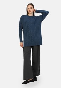 Overmaatse donkerblauwe gebreide trui met kabelpatronen, geribbelde zoom en lange mouwen, gecombineerd met grijze broek met wijde pijpen en zwarte schoenen.