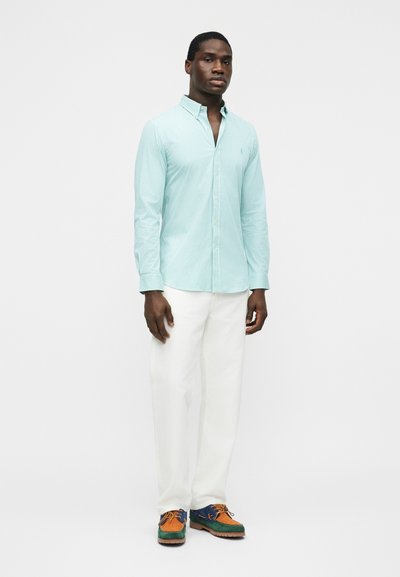 Egy férfi áll, világoskék csíkos ingben, fehér nadrágban és többszínű, fűzős cipőben, sima fehér háttér előtt.