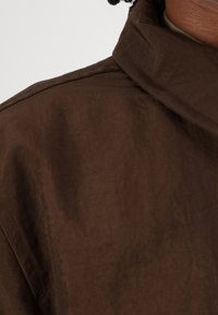 Veste marron en tissu texturé, avec un col montant et des détails de couture visibles le long des bords.
