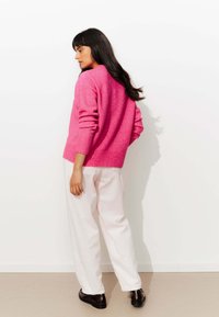 Pull en laine fuchsia avec des poignets et un ourlet côtelés, associé à un pantalon large blanc texturé et des chaussures noires, de dos par rapport au spectateur.