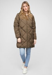 Veste matelassée verte olive avec une fermeture éclair à l'avant, une capuche et des poches latérales, associée à un jean bleu clair et des baskets blanches.