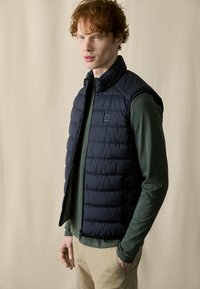 Dunkelblauer Steppwesten mit hohem Kragen und gestepptem Muster, getragen über einem grünen Langarmshirt mit Reißverschluss und Taschen.