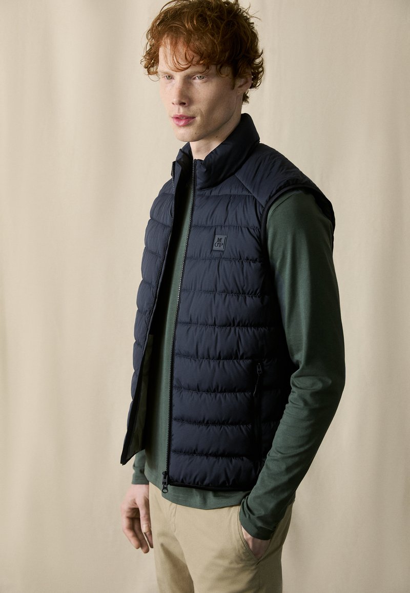 Dunkelblauer Steppwesten mit hohem Kragen und gestepptem Muster, getragen über einem grünen Langarmshirt mit Reißverschluss und Taschen.