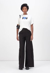 Witte T-shirt met zwart en blauwe grafische print, gecombineerd met hoge zwarte wijde broek met cargozakken. Zwarte schoenen en tas.