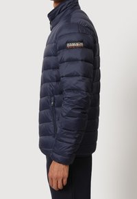 Uomo che indossa una giacca trapuntata blu navy con tasca laterale con zip e logo Napapijri Geographic sulla manica sinistra, in piedi di profilo.