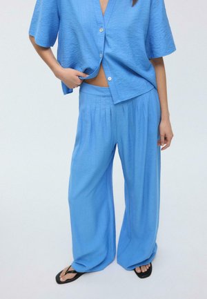 Žena oblečená v voľnom modrom dvojdielnom outfite s krátkym rukávom a zapínanou blúzkou a širokými nohavicami, doplnená čiernymi sandálmi so štvorcovými špičkami.
