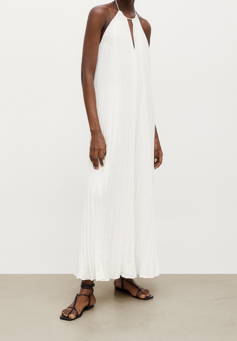 Personne à la peau foncée portant une robe maxi blanche plissée sans manches avec un col halter et des sandales plates noires, debout sur un sol clair.