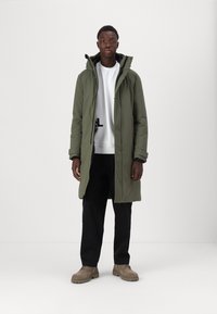Olivgrüner langer Parka mit einem Reißverschluss und Kapuze mit Kordelzug, über einem weißen Sweatshirt getragen, kombiniert mit schwarzen Hosen und beigen Schuhen.