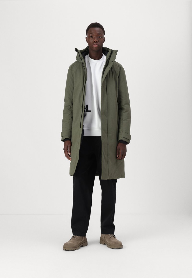 Olivgrüner langer Parka mit einem Reißverschluss und Kapuze mit Kordelzug, über einem weißen Sweatshirt getragen, kombiniert mit schwarzen Hosen und beigen Schuhen.