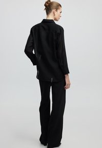 Camicia nera trasparente a maniche lunghe con colletto, dal taglio dritto e una leggera trasparenza, abbinata a pantaloni neri a gamba larga.